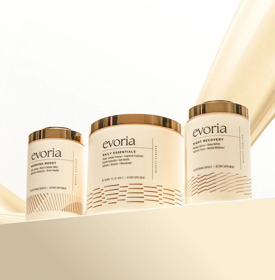 The complete Evoria supplement trio featuring Morning Boost capsules, Daily Essentials powder, and Night Recovery capsules. The three white and gold canisters are displayed together on an architectural cream shelf against a bright background.