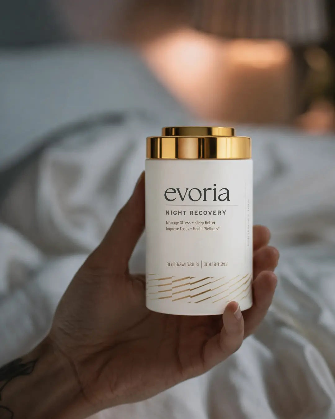 A hand holding a white and gold canister of Evoria Night Recovery supplement against a softly lit, blurred bedroom background.
