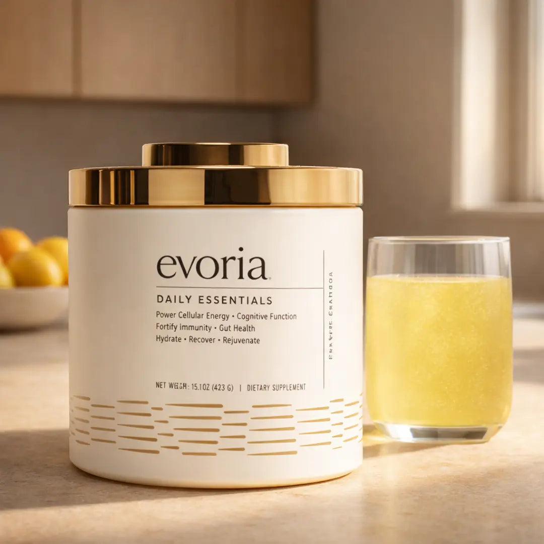 A white and gold canister of Evoria Daily Essentials dietary supplement next to a glass of the prepared yellow drink on a sunlit kitchen counter.